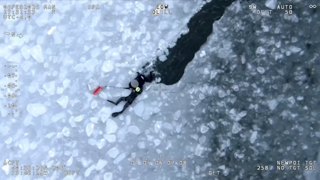 An aerial view from a drone shows a diver in a black wetsuit with a red flotation device lying on a broken ice sheet.